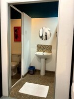 Un baño pequeño con un lavamanos blanco, un espejo redondo y un inodoro visible a través de una puerta abierta.