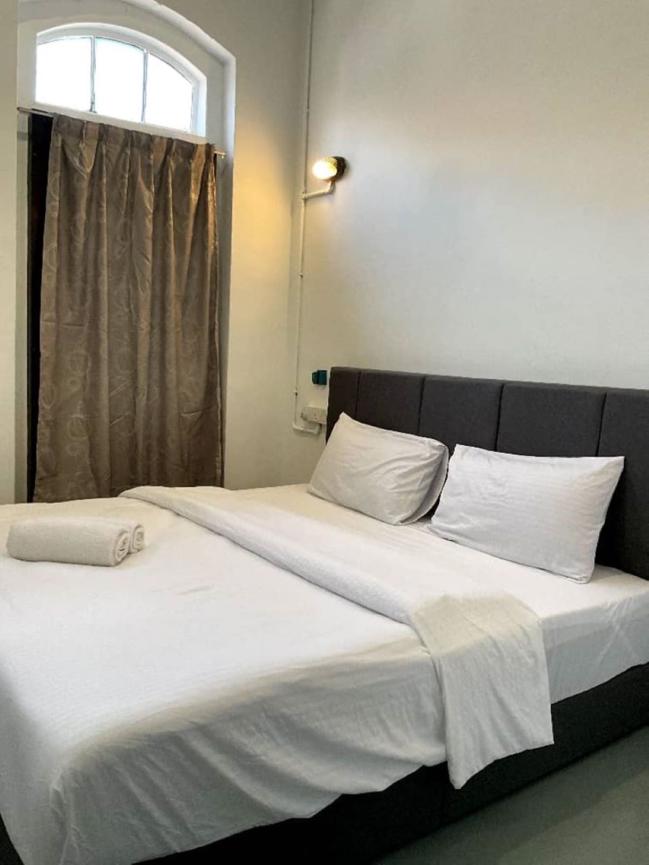 A neatly made bed with white linens and pillows, a dark headboard, and a window with curtains.