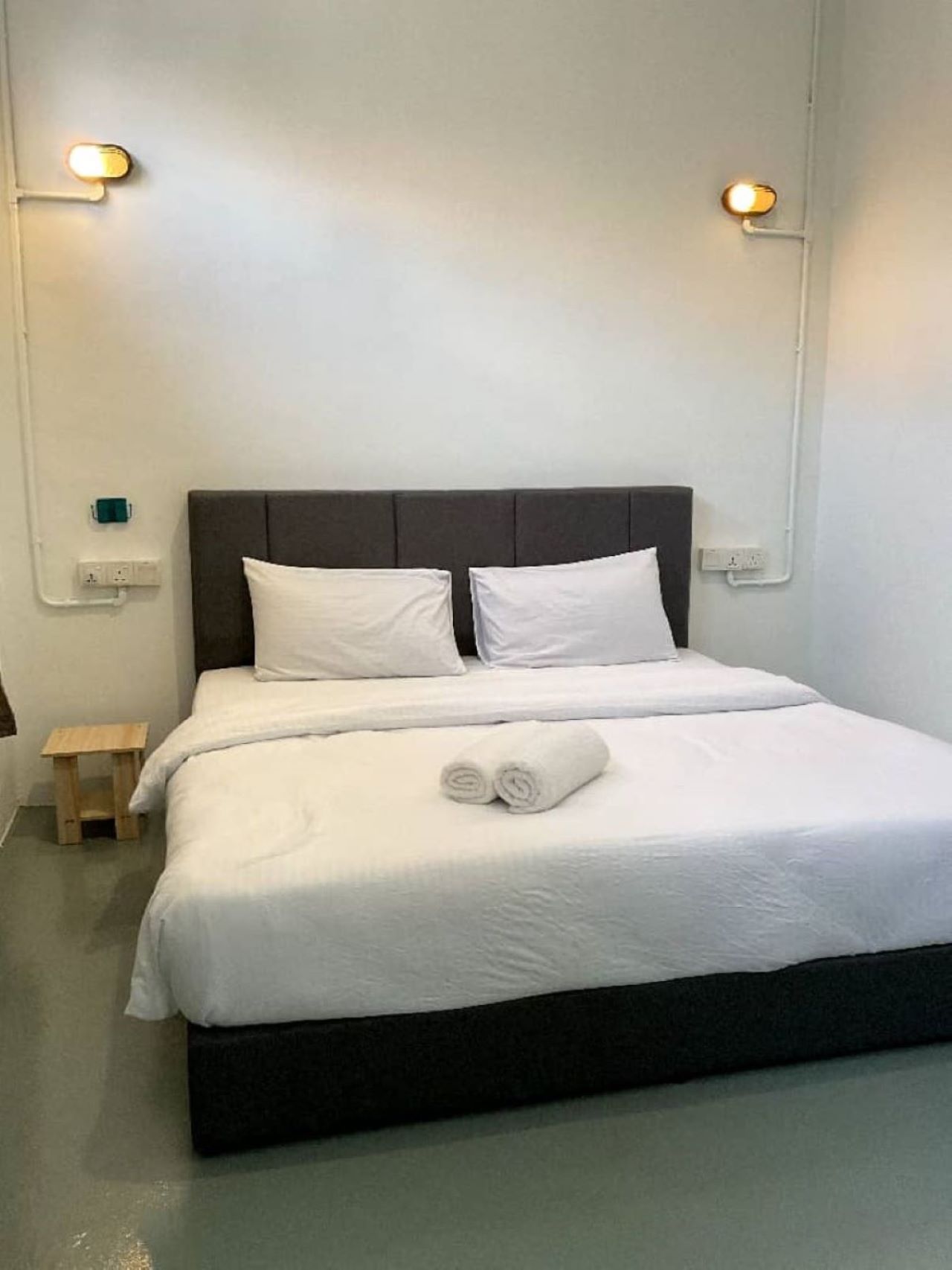 A neatly made bed with white linens and a rolled towel, a small wooden nightstand, and a wall-mounted light fixture.