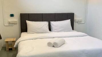 A neatly made bed with white linens and a rolled towel, a small wooden nightstand, and a wall-mounted light fixture.