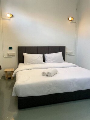 Cama bem arrumada com lençóis brancos, uma toalha enrolada, uma mesinha de cabeceira de madeira e um aplique na parede.