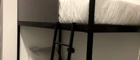 Una cama litera con escalera, ropa de cama blanca y un estante pequeño con una lámpara y un libro.