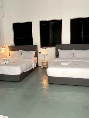 Quarto de hotel com duas camas, cada uma com roupa de cama e almofadas brancas, uma pequena mesa de cabeceira de madeira e uma luminária fixada na parede.