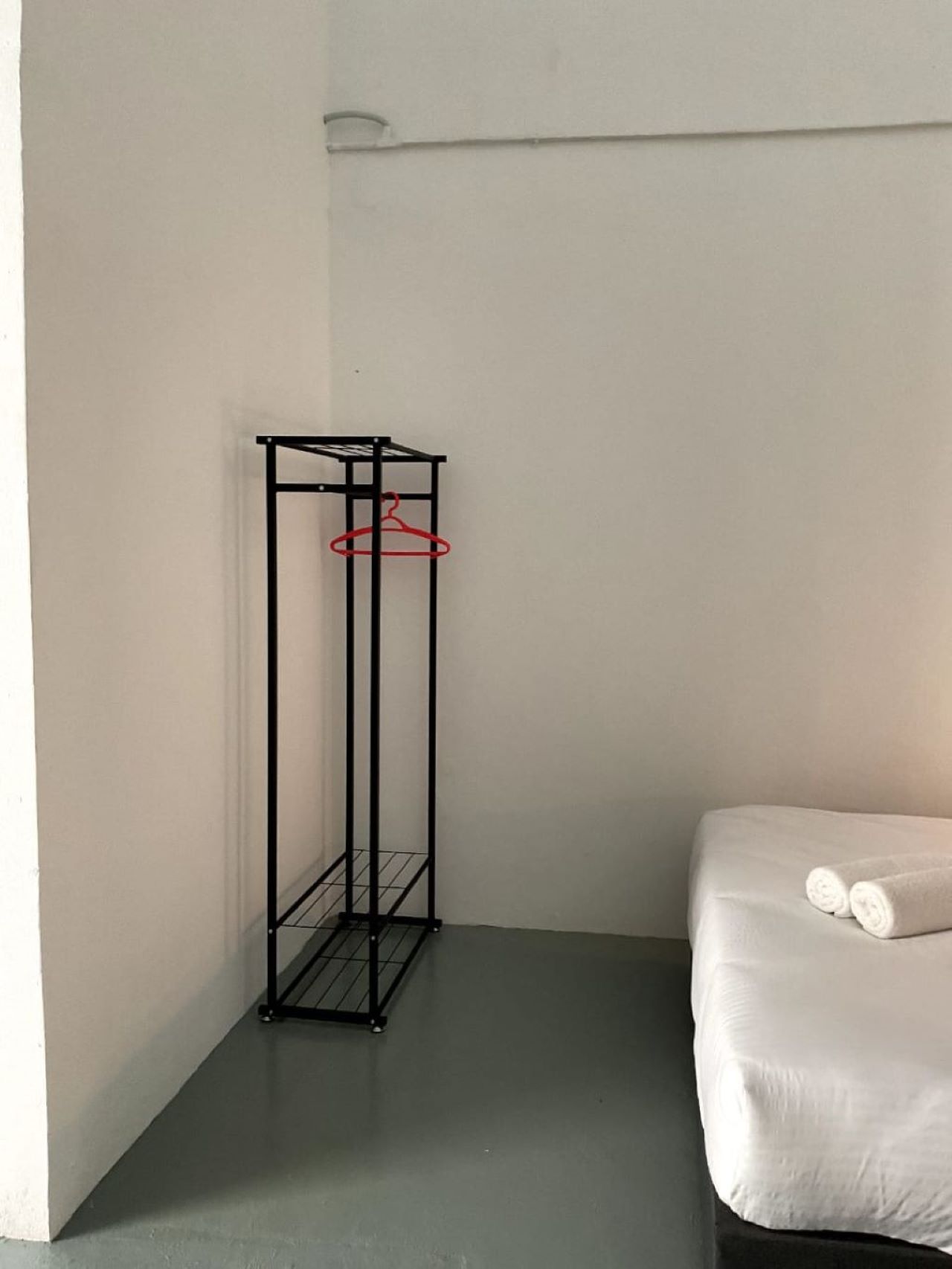 Una habitación pequeña y minimalista con una estructura metálica sosteniendo un perchero rojo, una cama blanca con toallas enrolladas y una pared lisa.