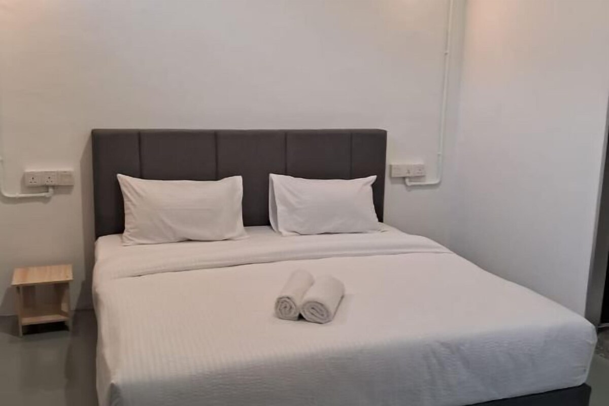 A neatly made bed with white bedding and pillows, a small wooden nightstand, and a rolled towel on the bed.