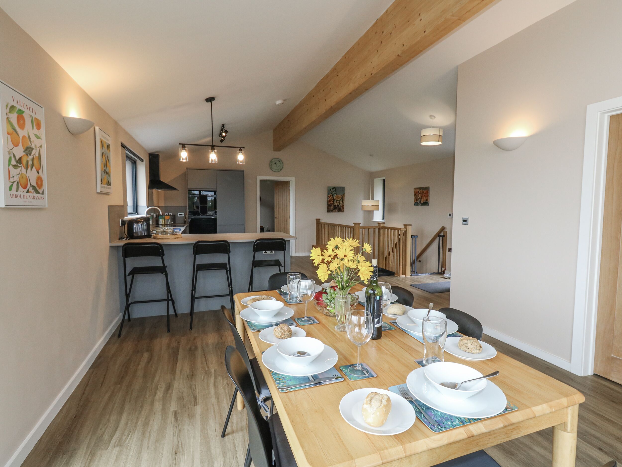 Un coin repas avec une table en bois dressée pour quatre personnes, une cuisine en arrière-plan et un escalier menant à un autre niveau.