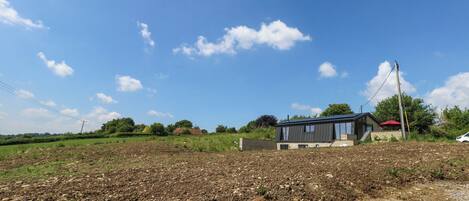 A modern house with a flat roof and large windows, surrounded by a rural landscape with open fields and scattered trees.
