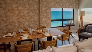 A dining area with a table set for six, chairs, a view of the ocean, a lamp, and a sofa.