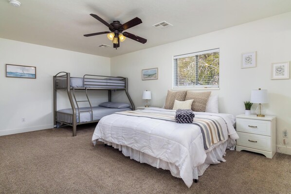 A bedroom with a bunk bed, a single bed, a ceiling fan, a window with blinds, and a bedside table with a lamp.