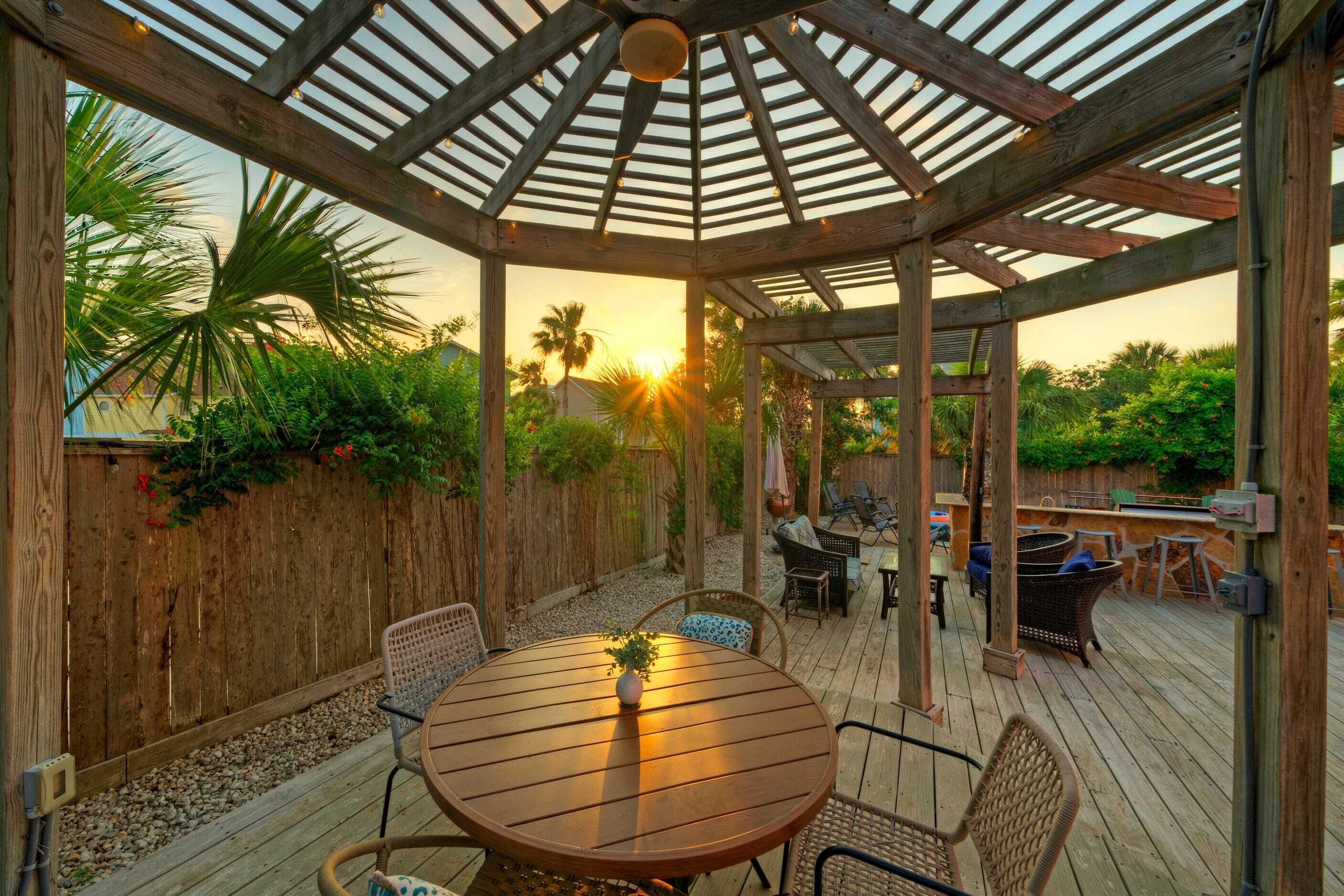 A wooden deck with a pergola, outdoor furniture, and a view of the sunset.