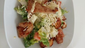 Un plato blanco con ensalada de lechuga, tomates, crutones y trozos de pollo a la parrilla.