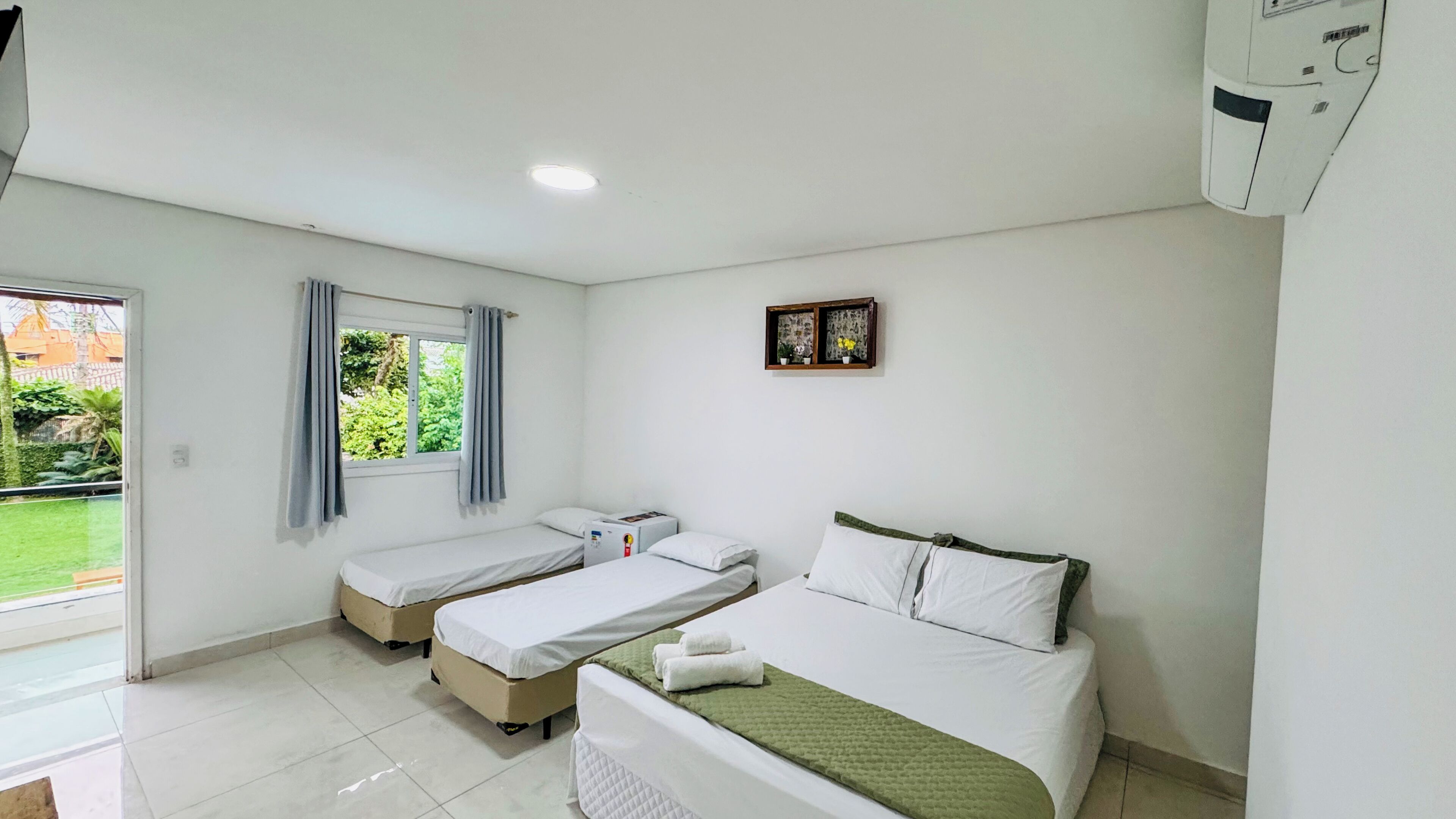 Quarto de hotel com duas camas de solteiro, janela com cortinas, prateleira pequena com objetos decorativos e ar condicionado no teto.