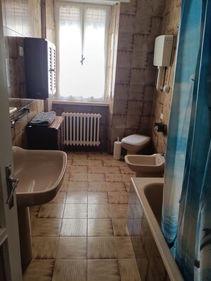 Ein Badezimmer mit einer Badewanne, zwei Waschbecken, einer Toilette, einem Fenster mit Vorhängen und einem Heizkörper.