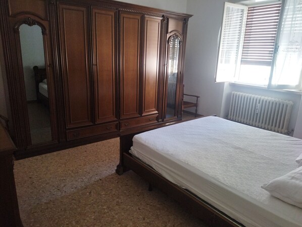 A bedroom with a wooden wardrobe, a bed with a white mattress, a radiator, and a window with blinds.