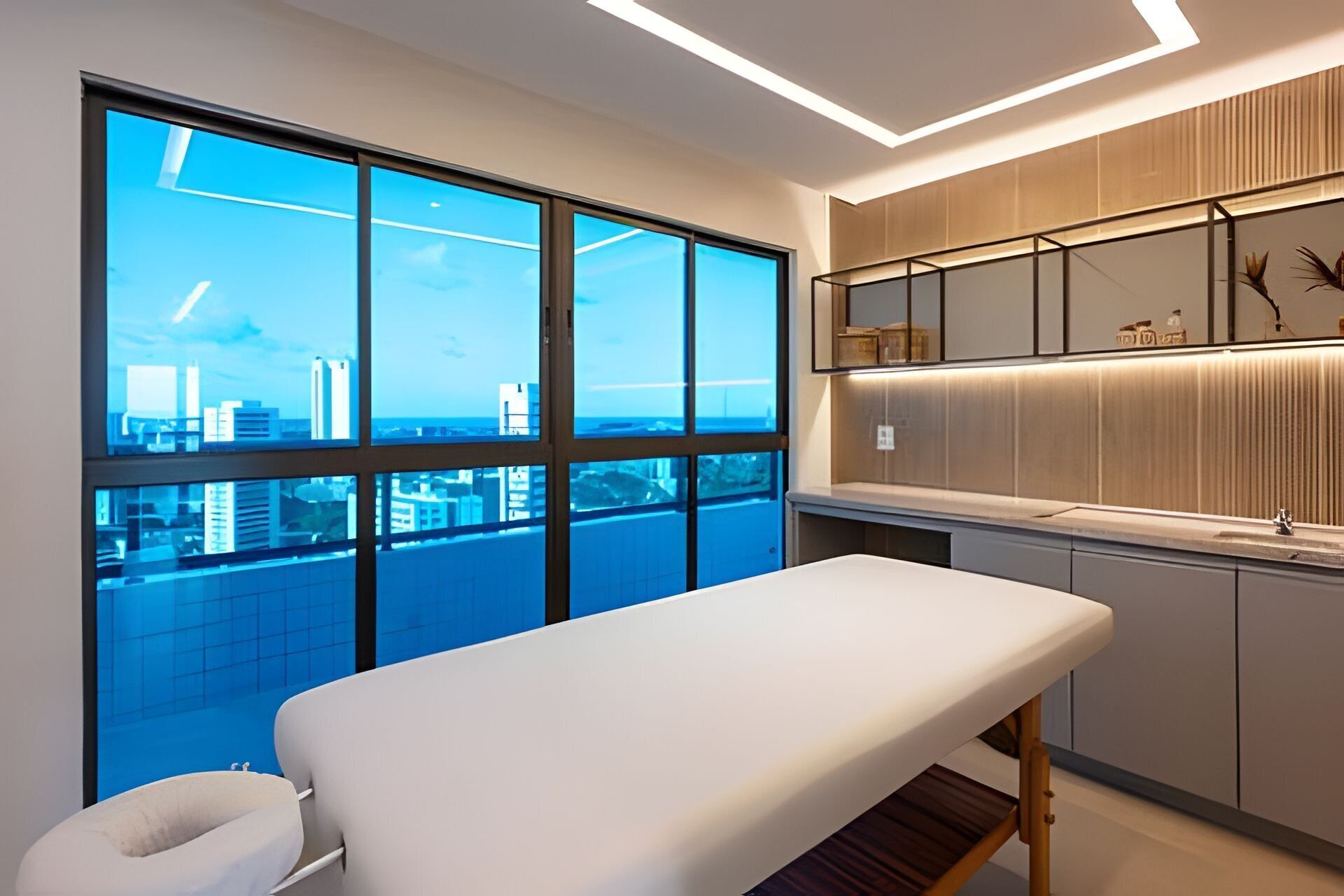Un moderno consultorio médico con un amplio ventanal que ofrece vista a la ciudad, una camilla blanca y un armario de madera con espejo y objetos decorativos.