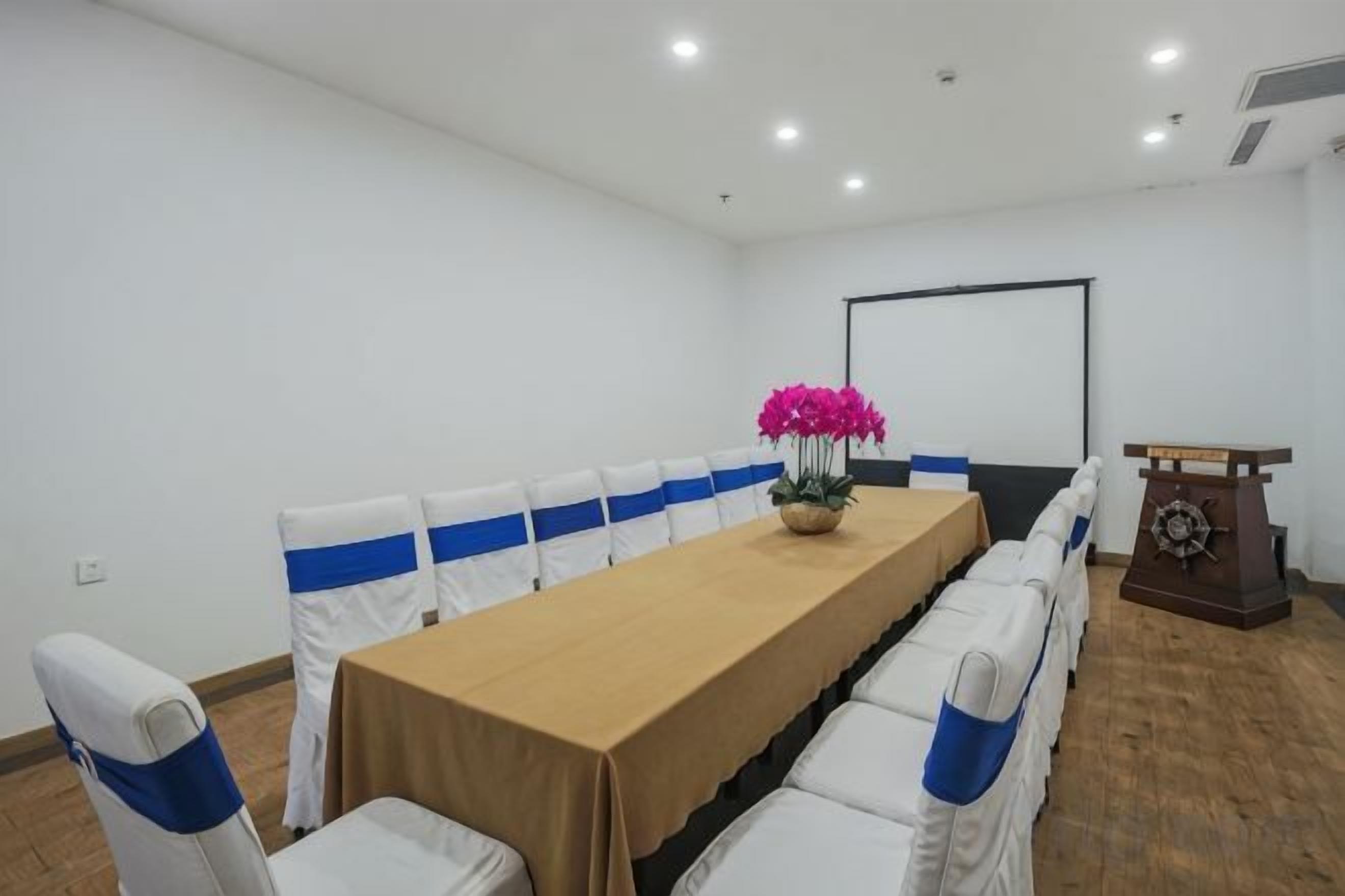 Un salón de conferencias con una larga mesa, sillas blancas con cintas azules, un atril y una pantalla de proyector.