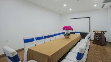 Un salón de conferencias con una larga mesa, sillas blancas con cintas azules, un atril y una pantalla de proyector.