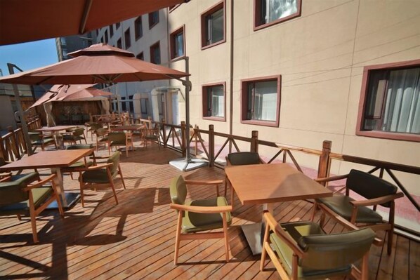 Un espace extérieur aménagé avec des tables et des chaises, des parasols, et la façade d’un bâtiment dotée de fenêtres.