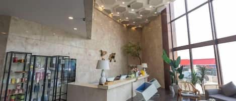 Um saguão de hotel com balcão de recepção, área de estar, janelas amplas e teto decorativo.