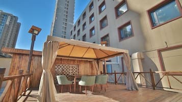 Ein Sitzbereich im Freien mit Holzterrasse, Markise und Blick auf hohe Gebäude.