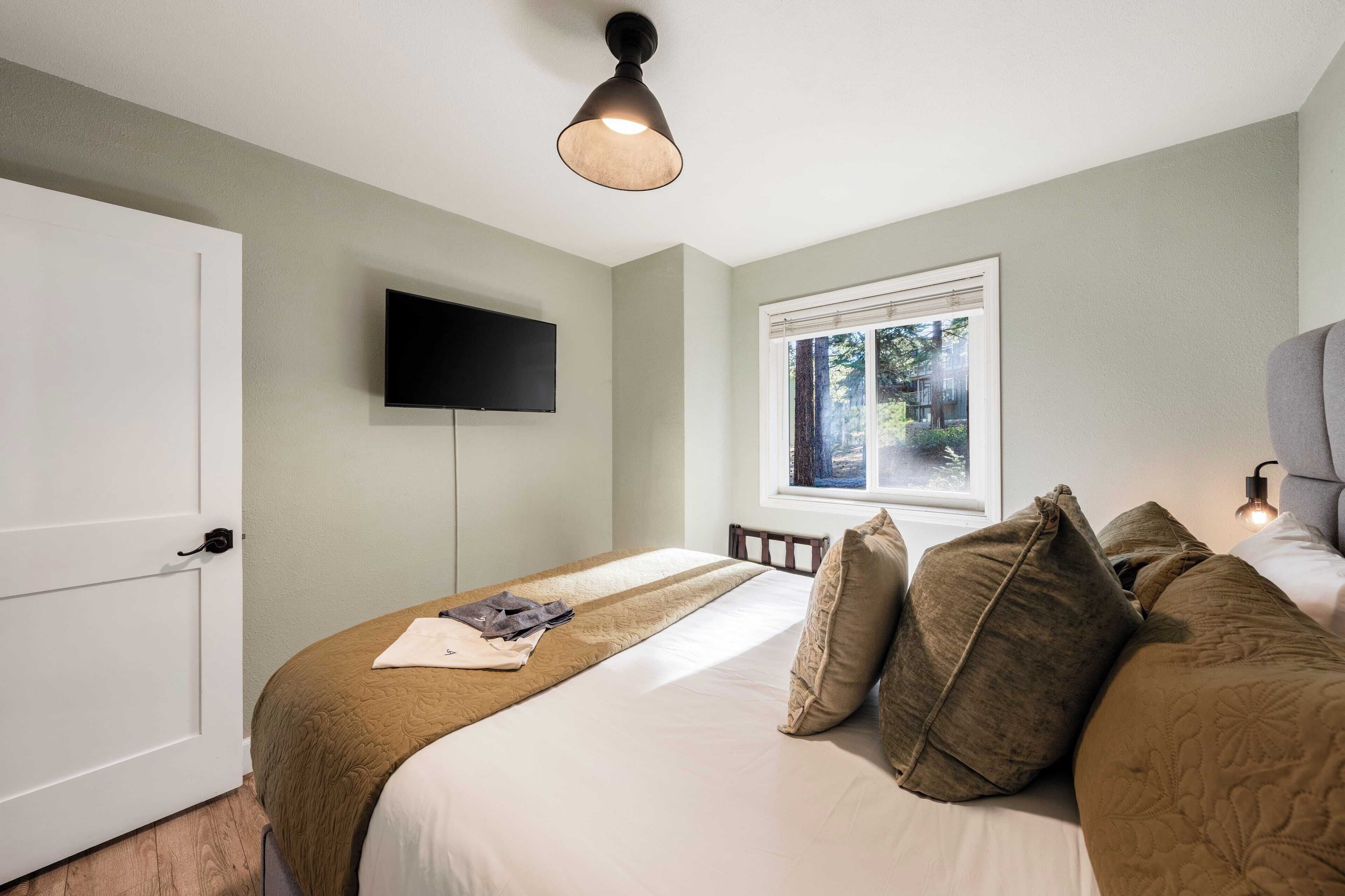 Une chambre à coucher avec un lit, un téléviseur fixé au mur, une fenêtre donnant sur des arbres et une porte.
