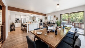 A modern kitchen with white cabinets, a wooden dining table, and a large window overlooking a deck. - Geraldine Drive by Avantstay Hot Tub, Views, Walk to Trails + Game Room (Incline Village)