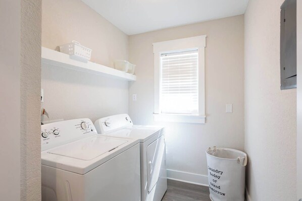 A laundry room with two white washers and dryers, a window with blinds, and a trash can with a message.