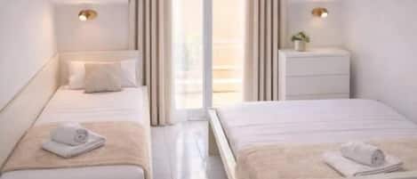 A modern hotel room with two beds, a small bedside table with a lamp, a window with curtains, and a view of the outside.