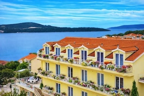 A multi-story building with balconies adorned with flowers, a view of a body of water, and a hilly landscape in the background. -  All Inclusive Villa Four Pearls   (Seget Donji)
