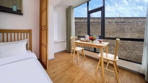 Une chambre d’hôtel avec un lit, une table à manger avec des chaises, une grande fenêtre donnant sur un mur de briques et un tableau accroché au mur.