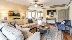 Ein geräumiges Wohnzimmer mit einem beige farbenen Sofa, einem blauen Sessel und einem Couchtisch. Es gibt einen Deckenventilator, einen an der Wand montierten Fernseher und ein großes Fenster mit Tageslicht.
