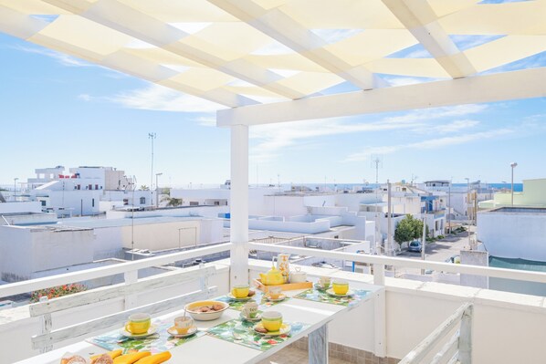 A rooftop terrace with a table set for breakfast, overlooking a residential area with white buildings and a clear sky.