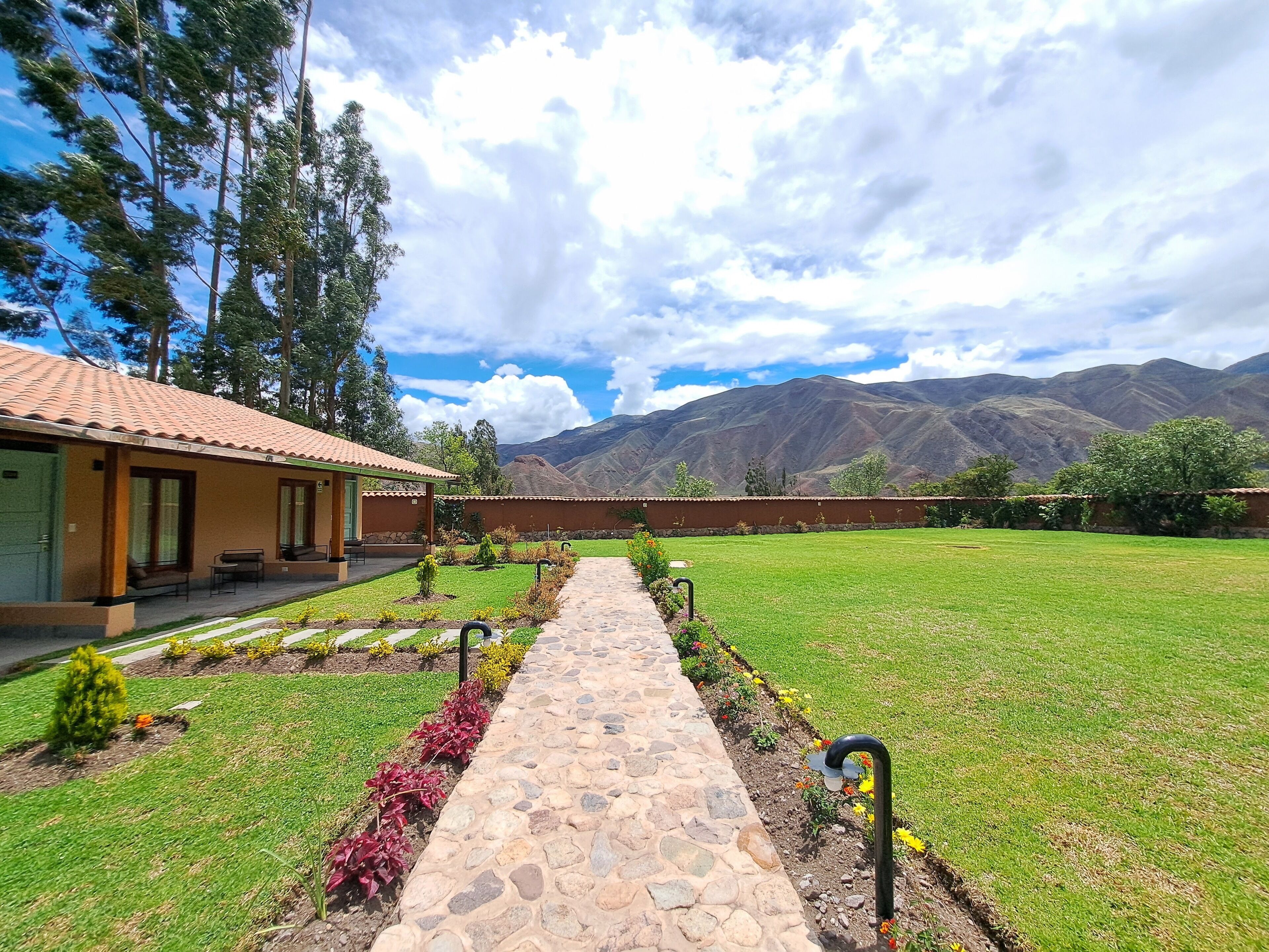 A pathway leading to a building with a tiled roof, surrounded by a well-maintained lawn and a mountain backdrop.