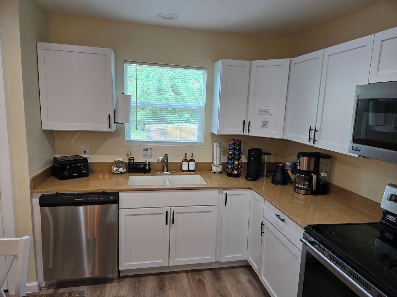 A modern kitchen with white cabinets, a stainless steel dishwasher, a built-in oven, a microwave, and a window providing natural light.
