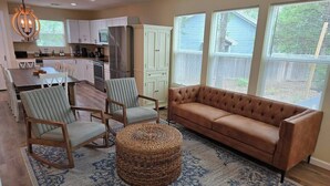 A spacious living room with a brown sofa, two armchairs, and a woven ottoman. A kitchen with white cabinets and a stainless steel refrigerator is in the background.