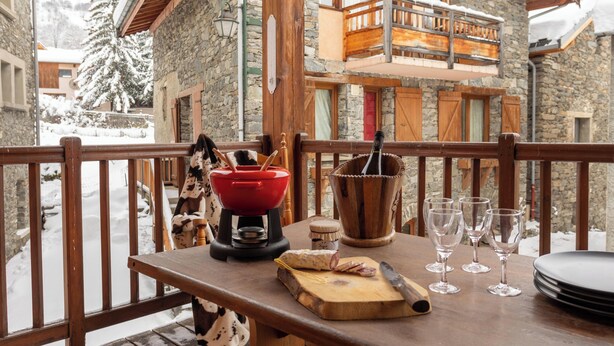 A wooden table with a red kettle, a cutting board with cheese, and wine glasses on a balcony overlooking a snowy village. - Chalet Laura, Ski & Authenticity (Les Belleville)