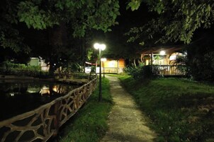 A nighttime pathway leading to a building with a covered patio area, surrounded by trees and a body of water.