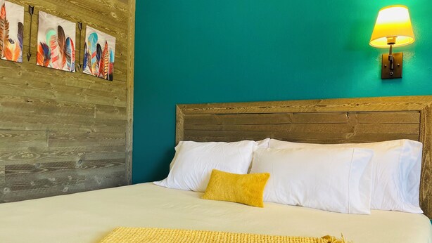 A neatly made bed with white and yellow bedding, a wooden headboard, and a wall-mounted lamp. - Historic Mora Inn and RV Park (Cleveland)