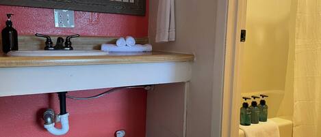 Un baño con una pared roja, un lavamanos blanco y un espejo. Hay toallas y artículos de aseo en el lavamanos, y una toalla colgada en la pared.