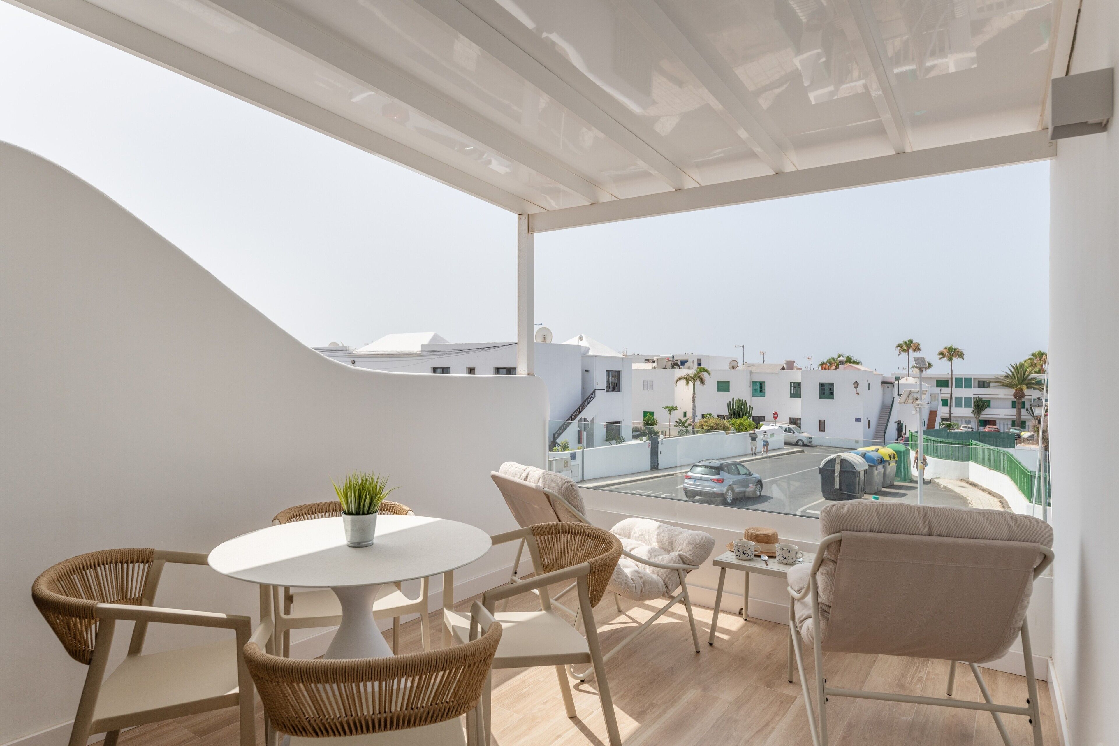 Un balcon avec une table blanche, des chaises en osier, un pot de fleurs et une vue sur une rue et des immeubles.