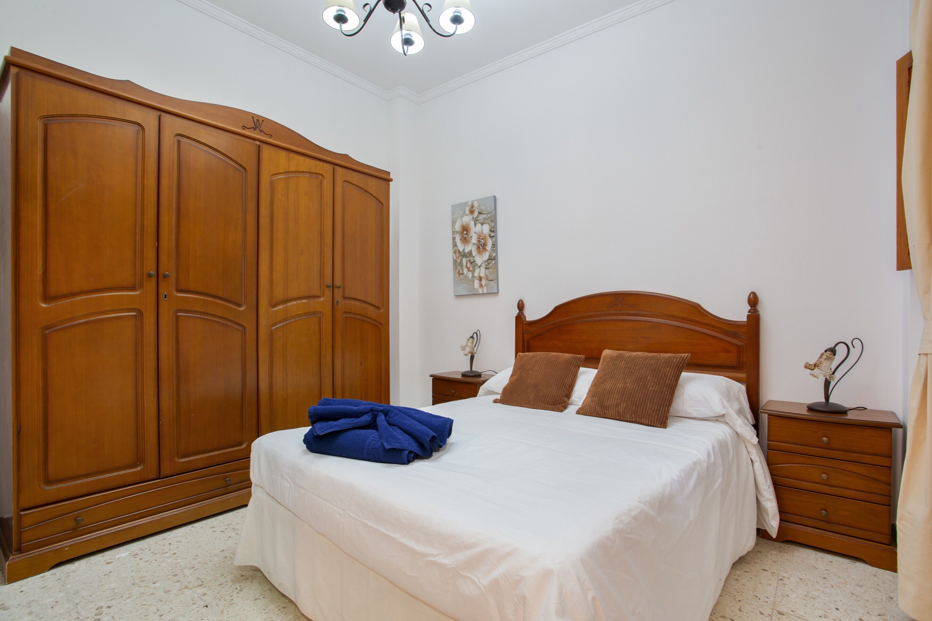 A bedroom with a large wooden wardrobe, a bed with white bedding, a nightstand with a lamp, and a wall-mounted artwork.