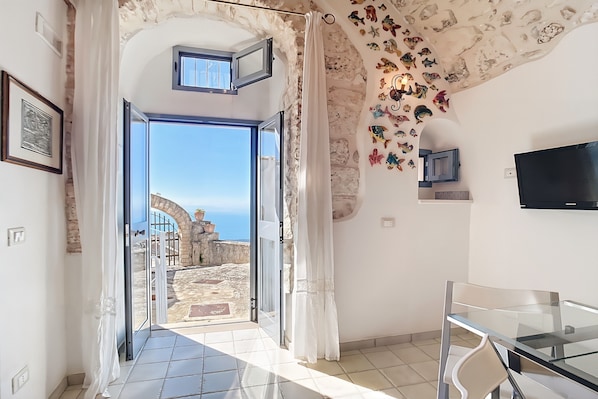 A room with a glass door leading to a view of a stone pathway and sea, a TV mounted on the wall, and a dining area with a glass table and chairs.