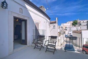 Un balcon avec deux chaises en bois, un bâtiment blanc avec une porte, et une vue sur une rue bordée de voitures et d’immeubles.
