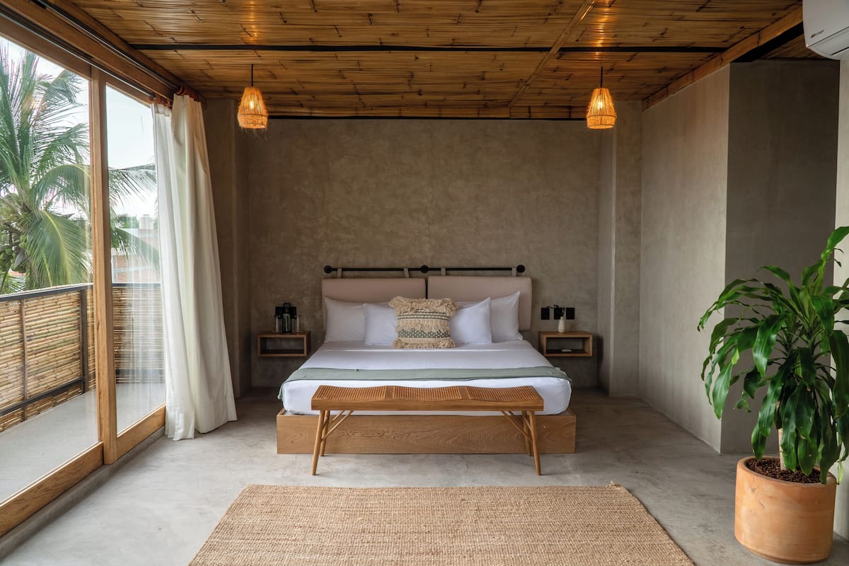 Una camera da letto con un letto grande, una testiera in legno, due comodini, una panca in vimini, un'ampia finestra con tende e una pianta in vaso.