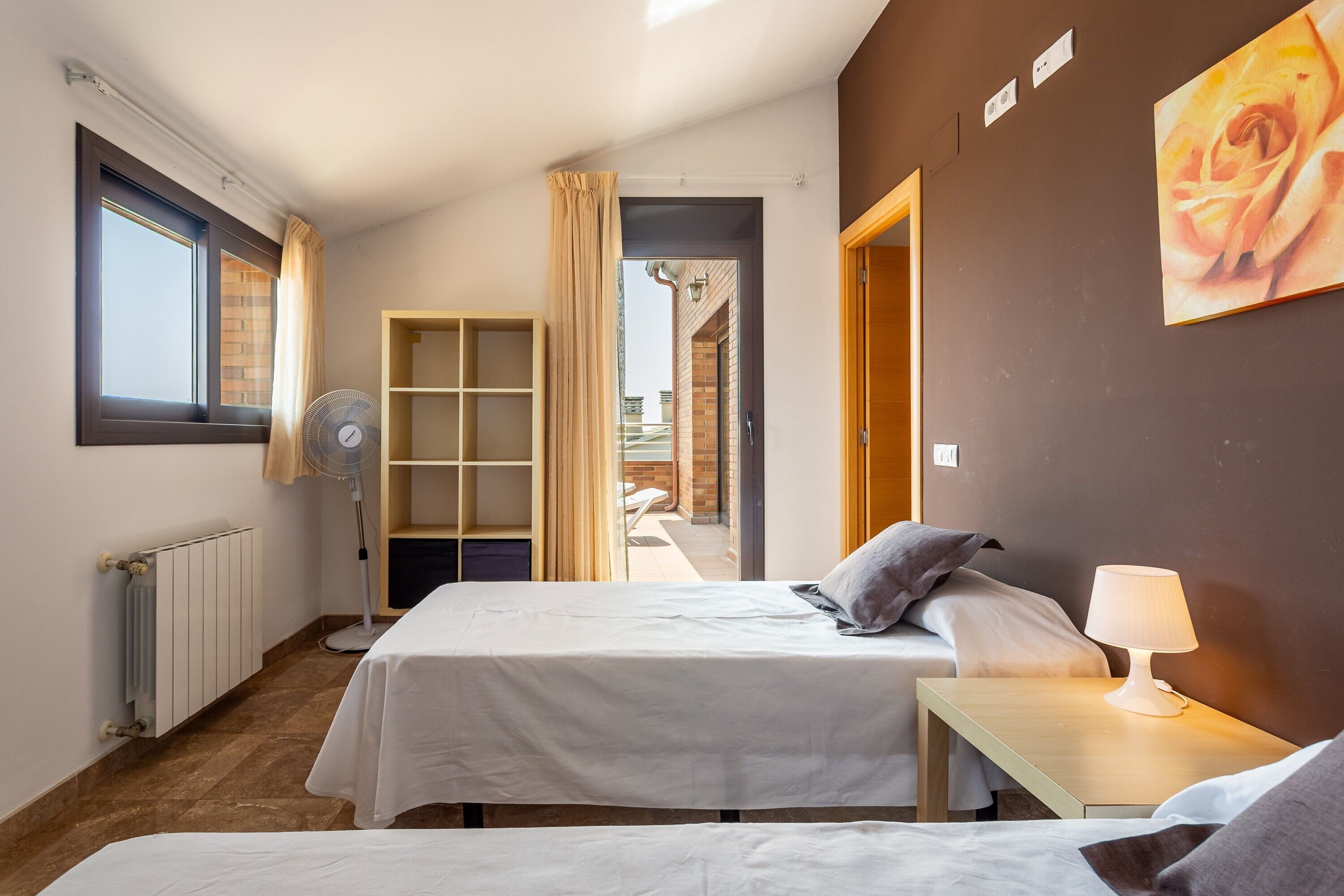 A hotel room with two beds, a bedside table with a lamp, a standing fan, a radiator, and a view of the outside.