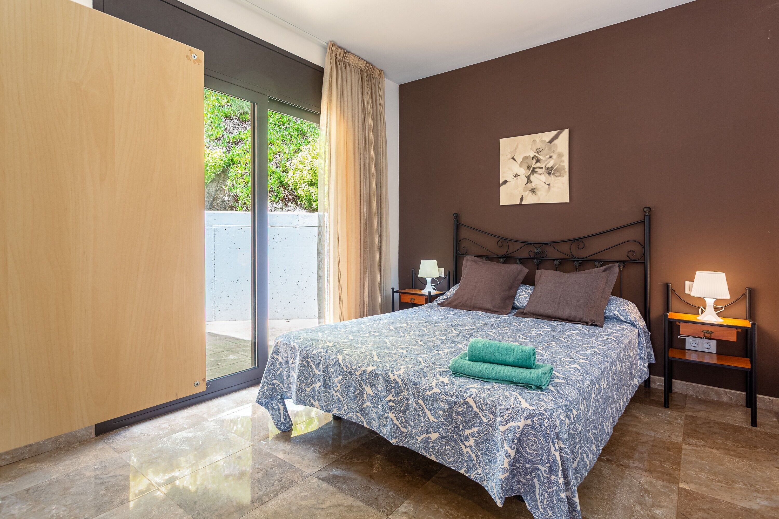A bedroom with a bed, a nightstand, a lamp, a window with curtains, and a wall-mounted artwork.