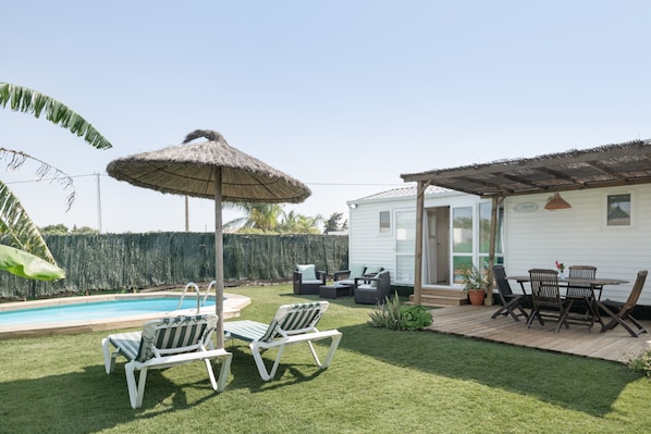 Um quintal com piscina, espreguiçadeiras, uma mesa e cadeiras, e um guarda-sol de palha.