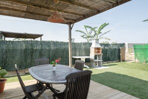 En overdækket terrasse med et rundt bord og stole, en hængende lysekrone, en murstensovn og et grønt hegn.