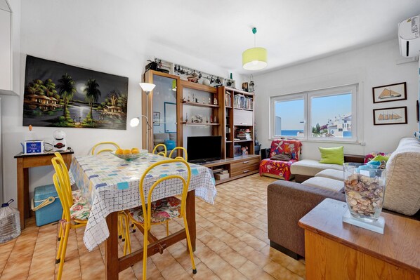 A living room with a dining table, chairs, a TV, a bookshelf, and a view of the sea through the windows.