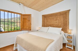 A bedroom with a large bed, a window with a view of greenery, a wooden ceiling, and a woven tapestry.